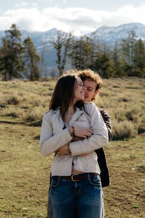 Couple embracing in a mountain meadow, woman in white jacket and jeans, snow-capped peaks in background.