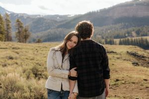 A couple embraces in a scenic mountain meadow, with pine trees and snow-capped peaks in the background.