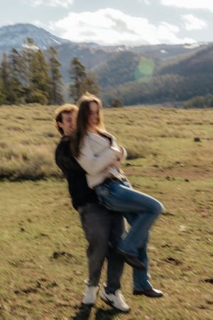 Couple joyfully spinning in a mountain meadow with snow-capped peaks and pine trees in the background.