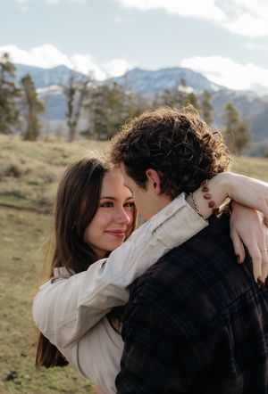 A couple embraces outdoors with snow-capped mountains in the background, the woman smiling warmly over the man's shoulder.