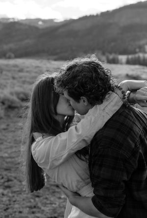 Black and white photo of a couple sharing a romantic kiss in a mountain meadow landscape.