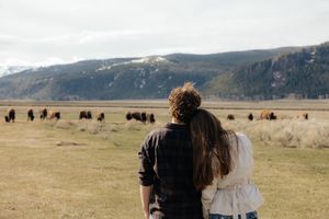 Couple standing together in a field watching bison graze with snow-capped mountains in the background.
