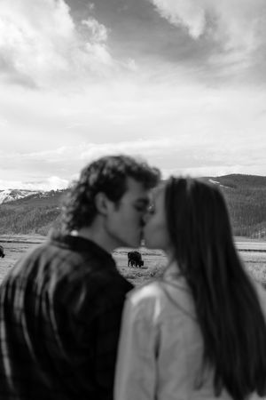 Black and white photo of couple kissing with bison grazing in snowy mountain landscape behind them.