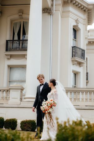 A bride and groom stand together in front of a grand, historic building, holding a bouquet of flowers.