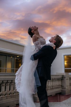 A bride and groom embrace on a balcony with a colorful sunset sky in the background.