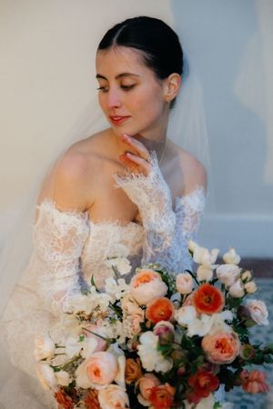 A bride in a lace wedding dress holds a bouquet of flowers and looks thoughtfully away from the camera.