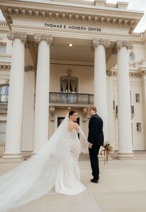 A bride in a flowing white dress and a groom in a dark suit stand together in front of a grand, columned building.