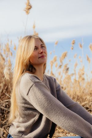 A young woman with long blonde hair wearing a gray sweater sits in a field of tall grass.
