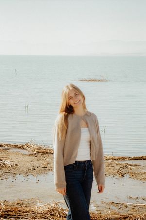 A young woman with blonde hair stands on a beach, smiling and wearing a beige jacket.