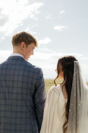 A bride and groom stand together outdoors, viewed from behind, with the bride wearing a pearl-adorned veil under a cloudy sky.