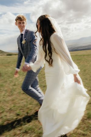 Bride in white dress and veil holds grooms hand running through green field, groom smiles in blue plaid suit outdoors.