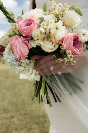 Bridal bouquet of pink roses, white blooms, and daisies held by bride in white dress outdoors.