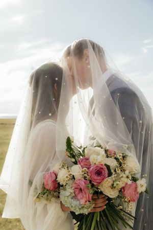 Bride and groom kissing under a sheer veil outdoors, bride holding a bouquet of pink and white roses.