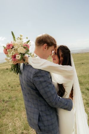 Bride and groom embrace in a green field, she holds pink and white bouquet, wearing veil and white dress, he wears blue plaid suit.