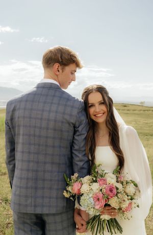 Bride in white gown holding pink and white bouquet leans on groom in plaid suit outdoors.