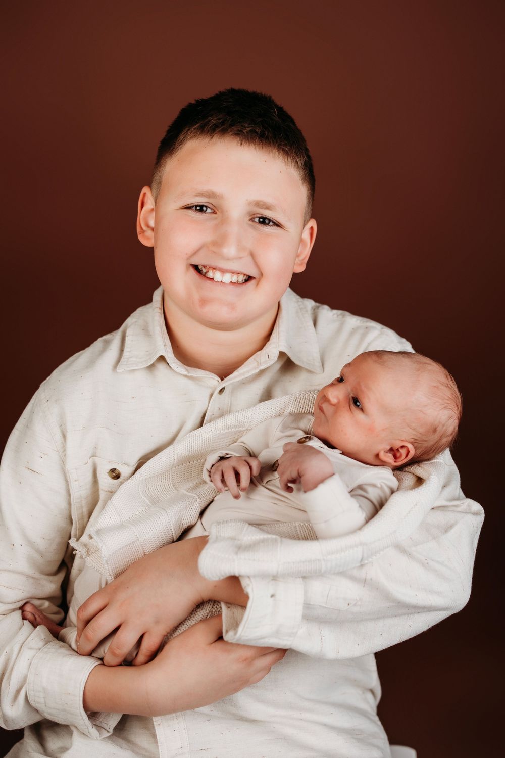 Two siblings in white clothing share tender moments against a dark brown background.