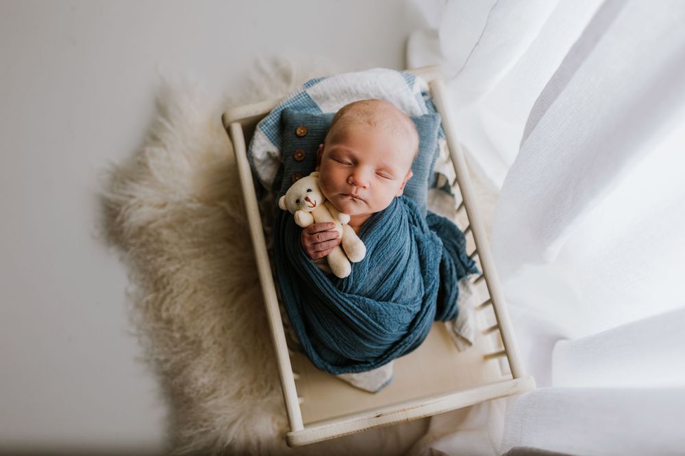Artistic newborn portrait with baby wrapped in teal fabric on wooden prop with natural lighting.