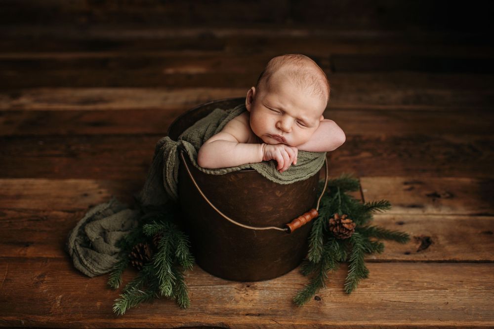 Baby rests peacefully in wooden bowl with evergreen branches on rustic wood surface.