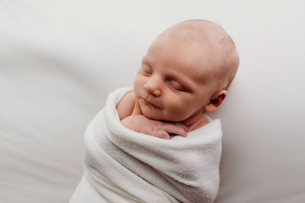 Sleeping newborn baby swaddled in white blanket against light background.