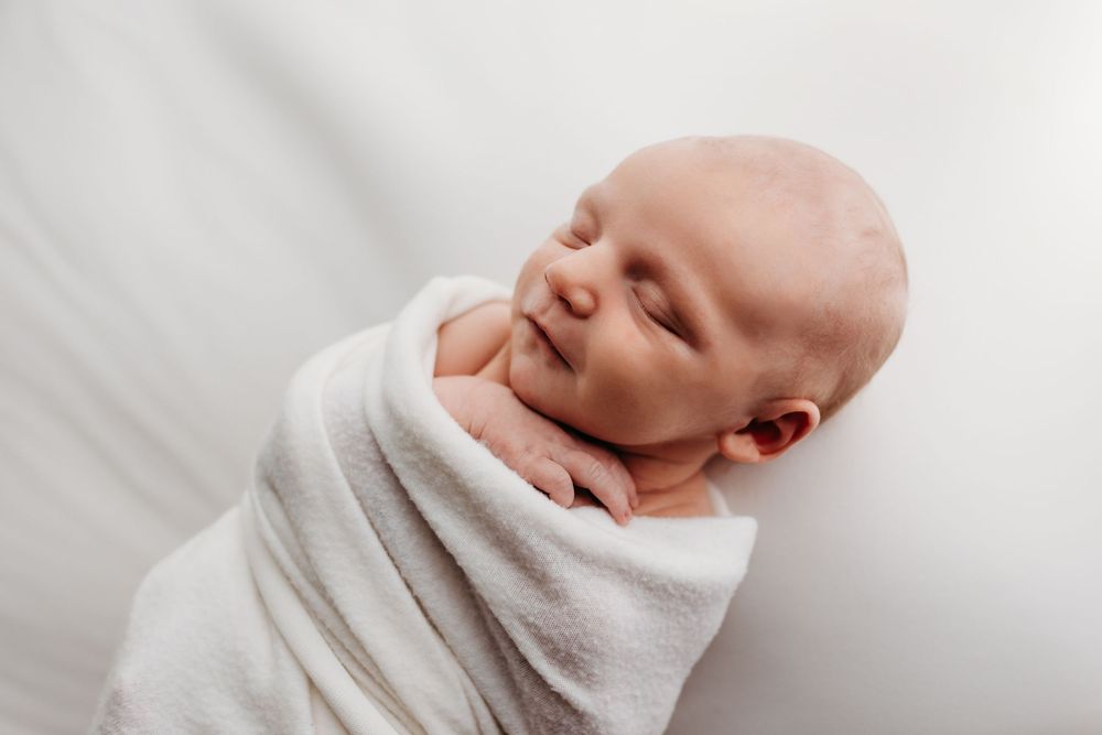 Minimalist newborn photography with baby wrapped in white against clean white background.