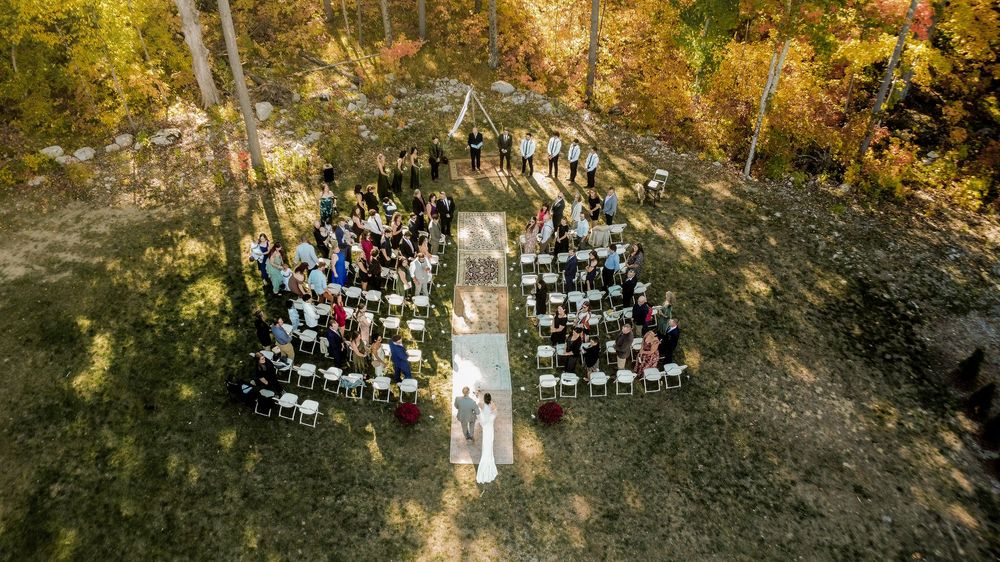 A panoramic view of an outdoor wedding ceremony with guests seated in a curved arrangement among autumn trees.