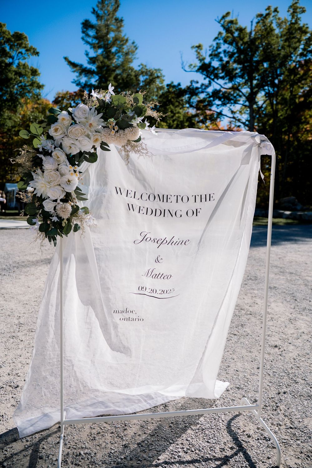 Three white fabric wedding welcome signs with elegant script text displayed outdoors with floral decorations.