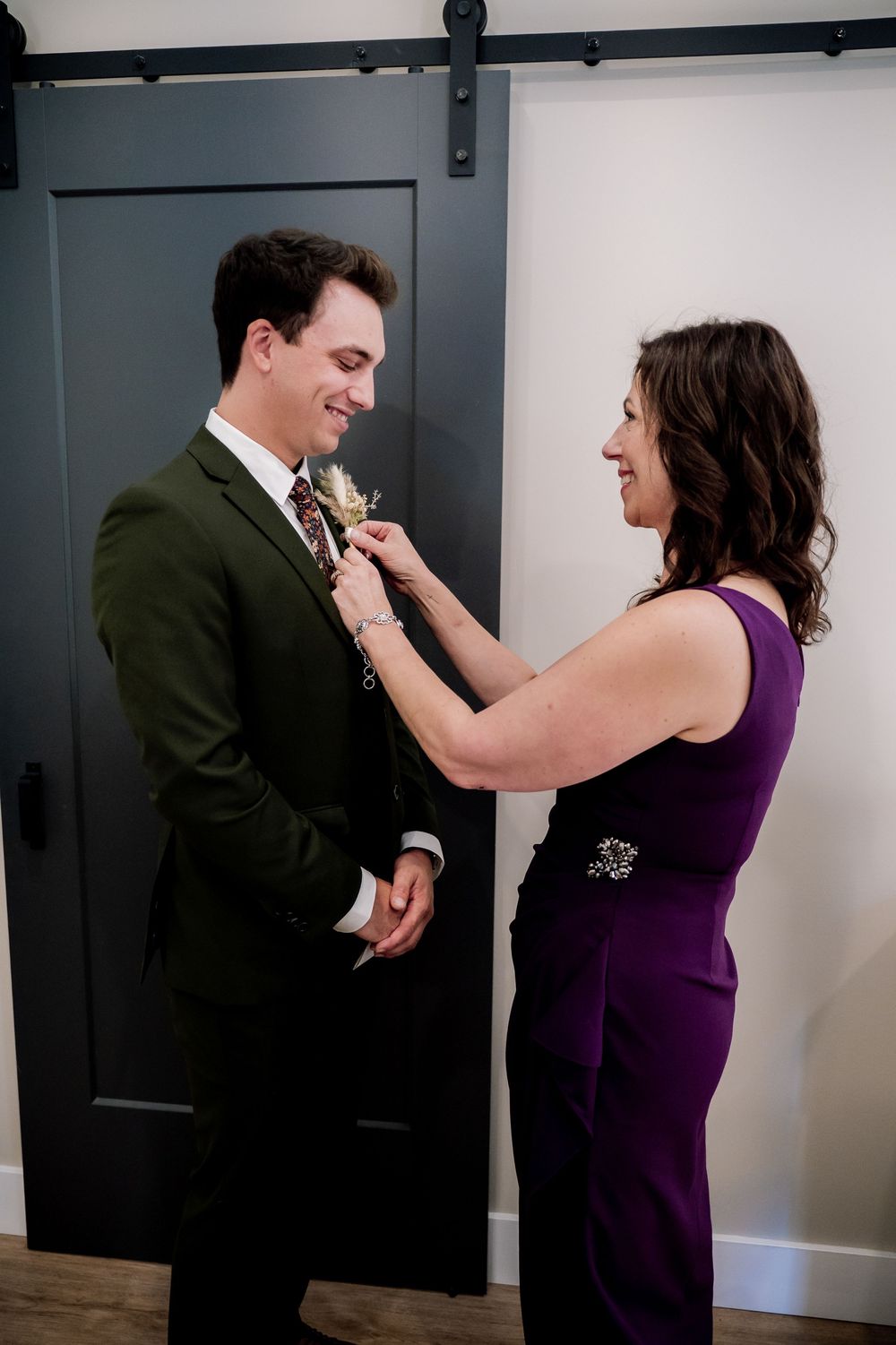 Two people sharing a tender moment while adjusting formal attire against a modern sliding barn door background.