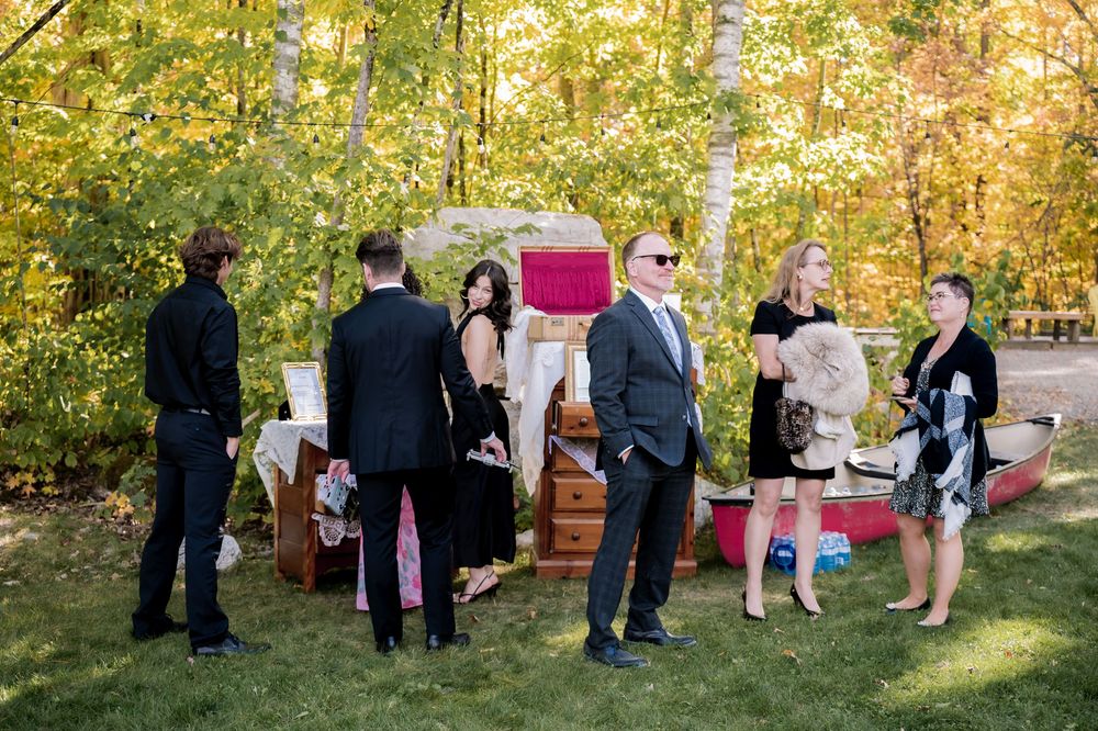 Wedding guests gather around a vintage camper trailer serving as a photo booth in an autumn outdoor setting.