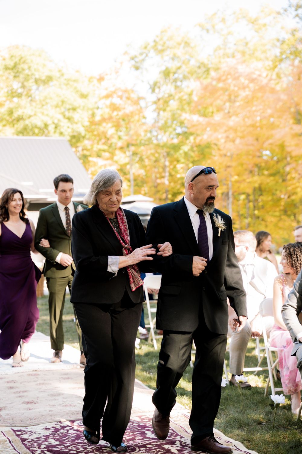 A wedding party dances in a line on a grassy lawn with autumn trees in the background.