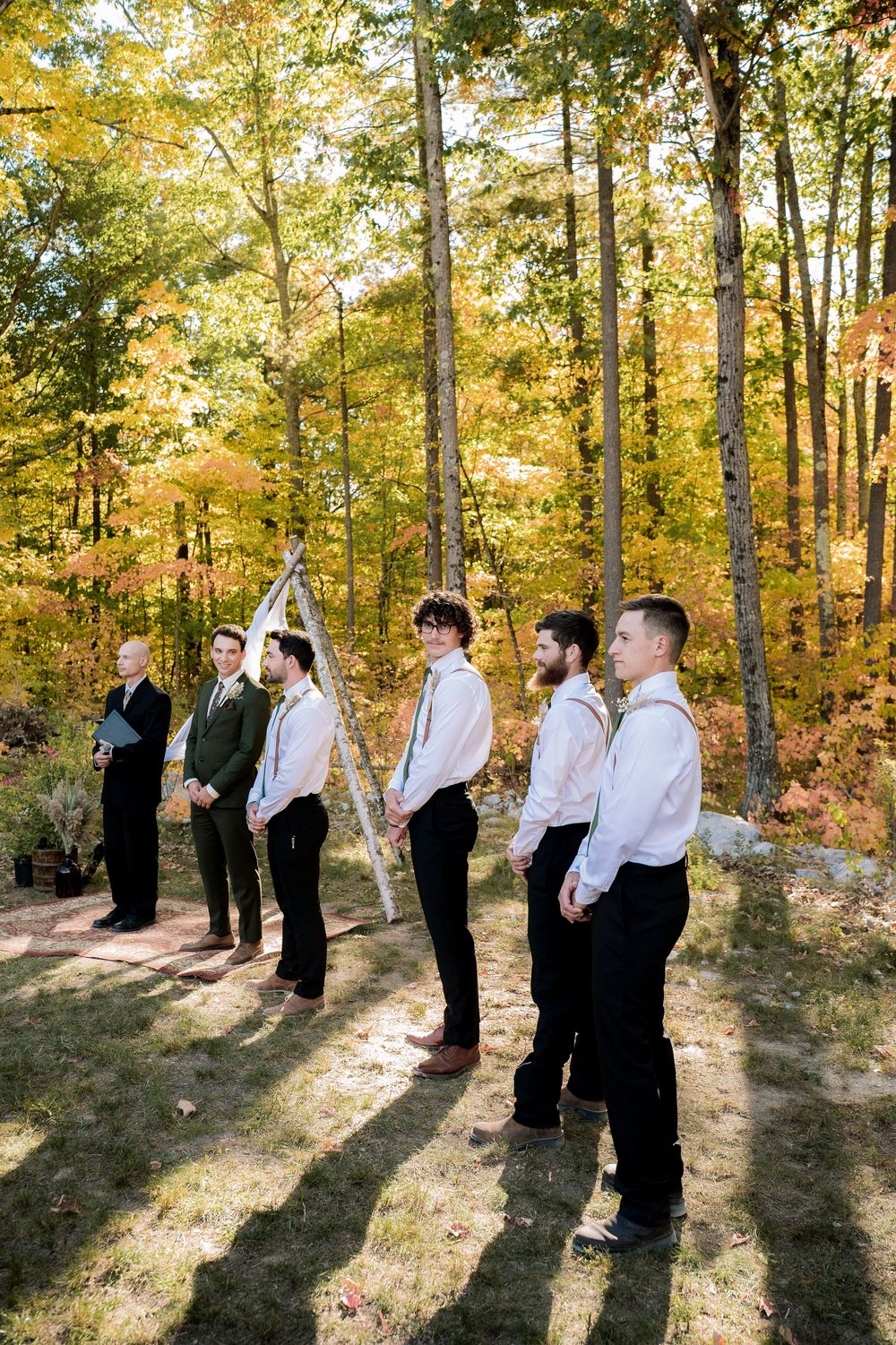 Wedding party members in formal attire gather in a sunlit autumn forest setting.