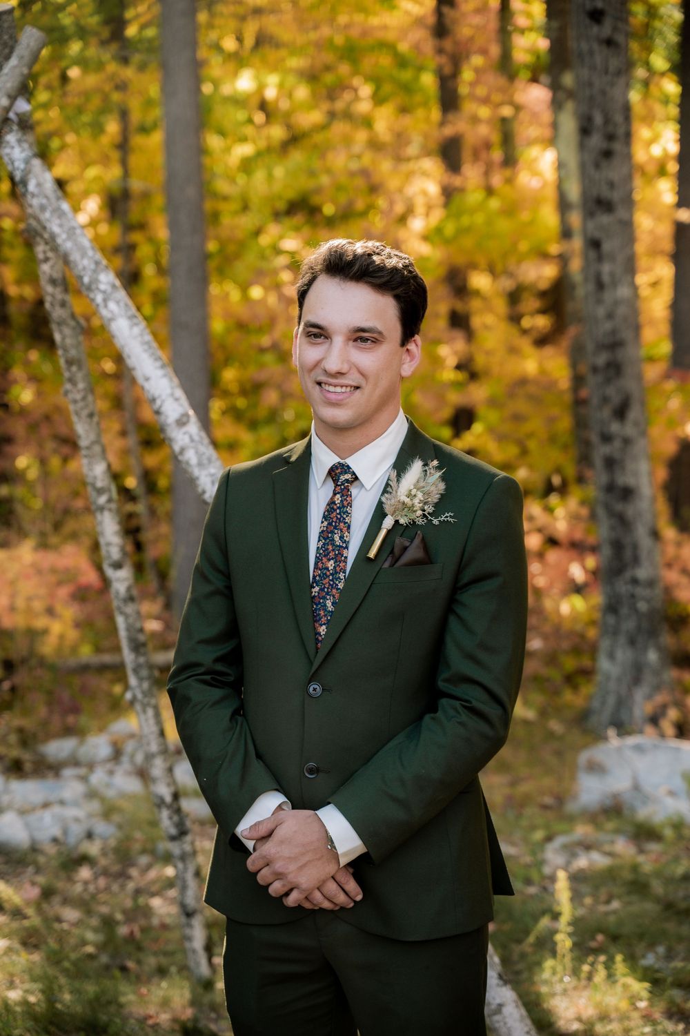 A groom in a dark green suit with a floral tie poses outdoors against a backdrop of autumn trees.