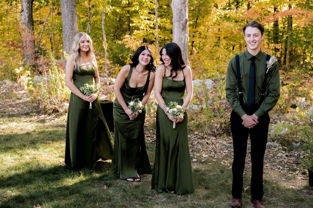 A group in formal black attire poses for photos in a sunlit autumn forest with golden leaves in the background.