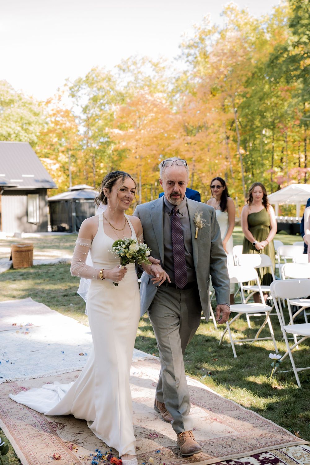 A wedding ceremony in a backyard setting with autumn trees and white folding chairs on a sunny fall day.
