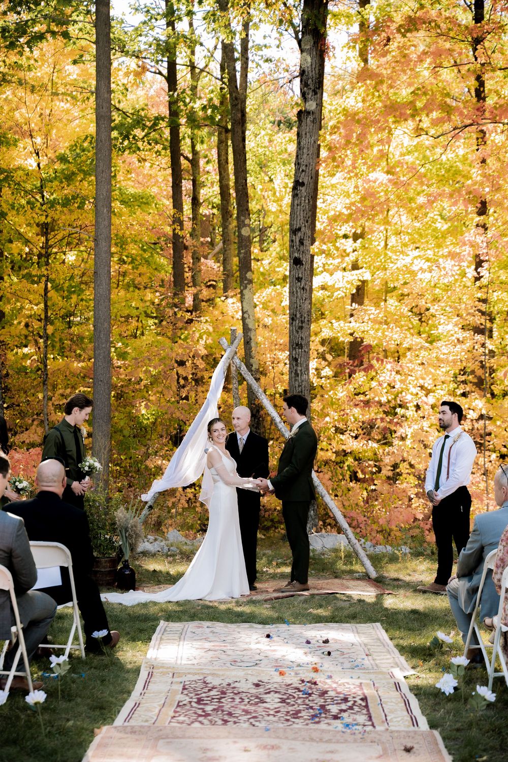 Outdoor wedding ceremony in progress with guests seated on chairs and a decorative rug aisle runner.