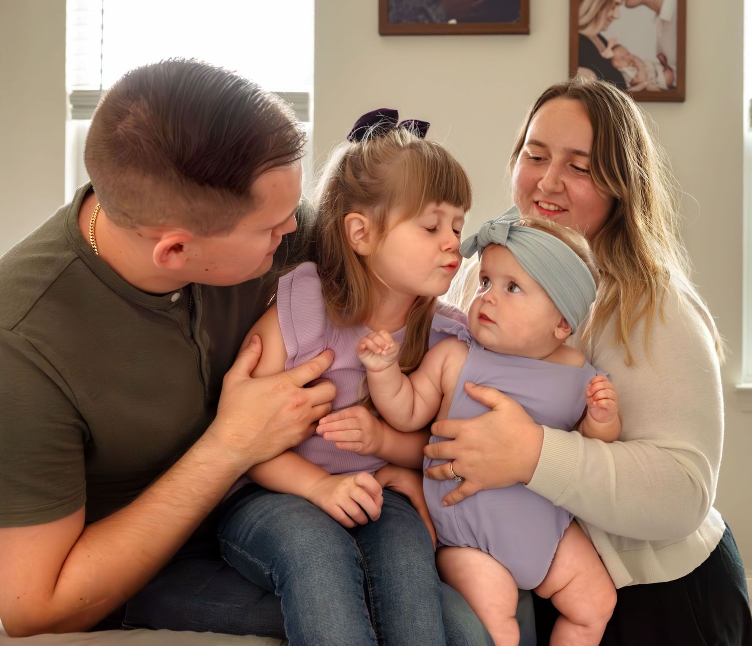 Family of four snuggling close together on the end of the bed. The toddler tries to kiss her baby sister. Doughman Photography