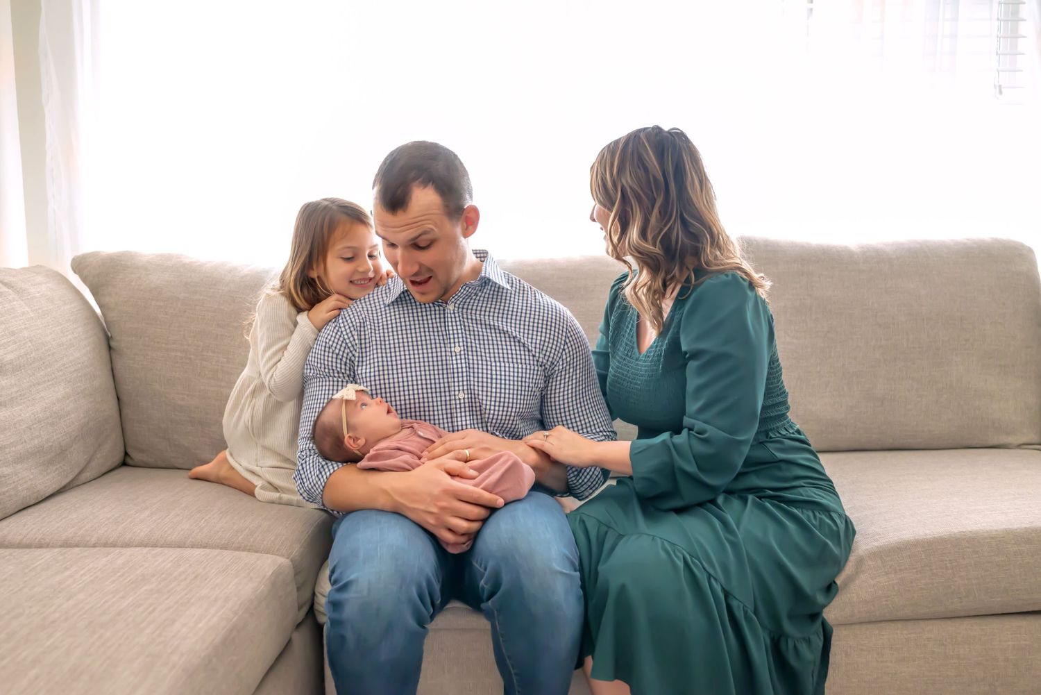 Oldest daughter snuggles dad, smiling at newborn sister in dad's arms. Mom is reaching behind dad for the middle child. Doughman Photography
