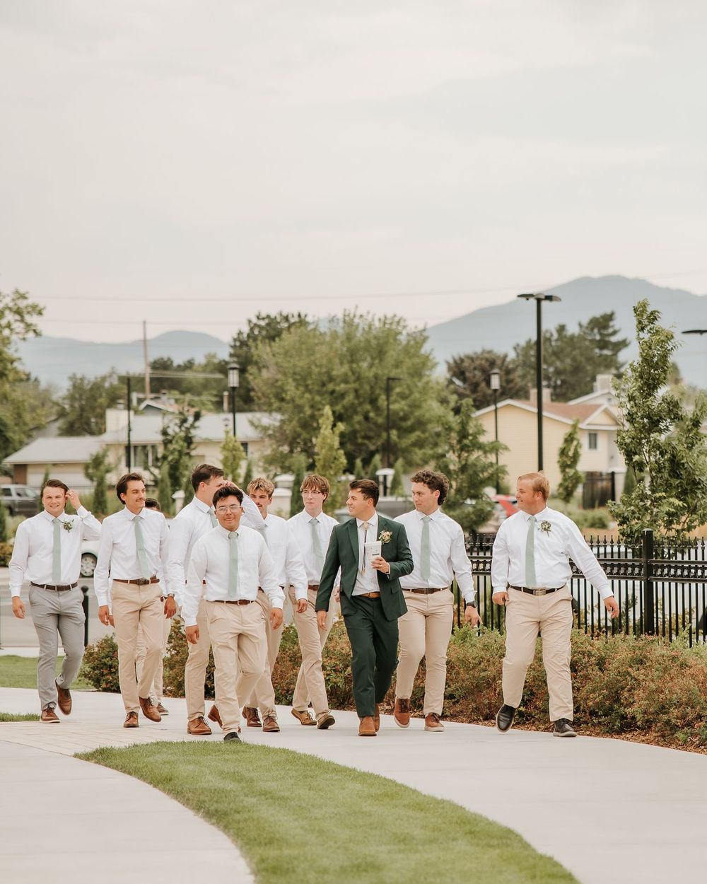 Groomsmen in light colored attire walking together on pathway with mountains in background with their dirty soda in hand.