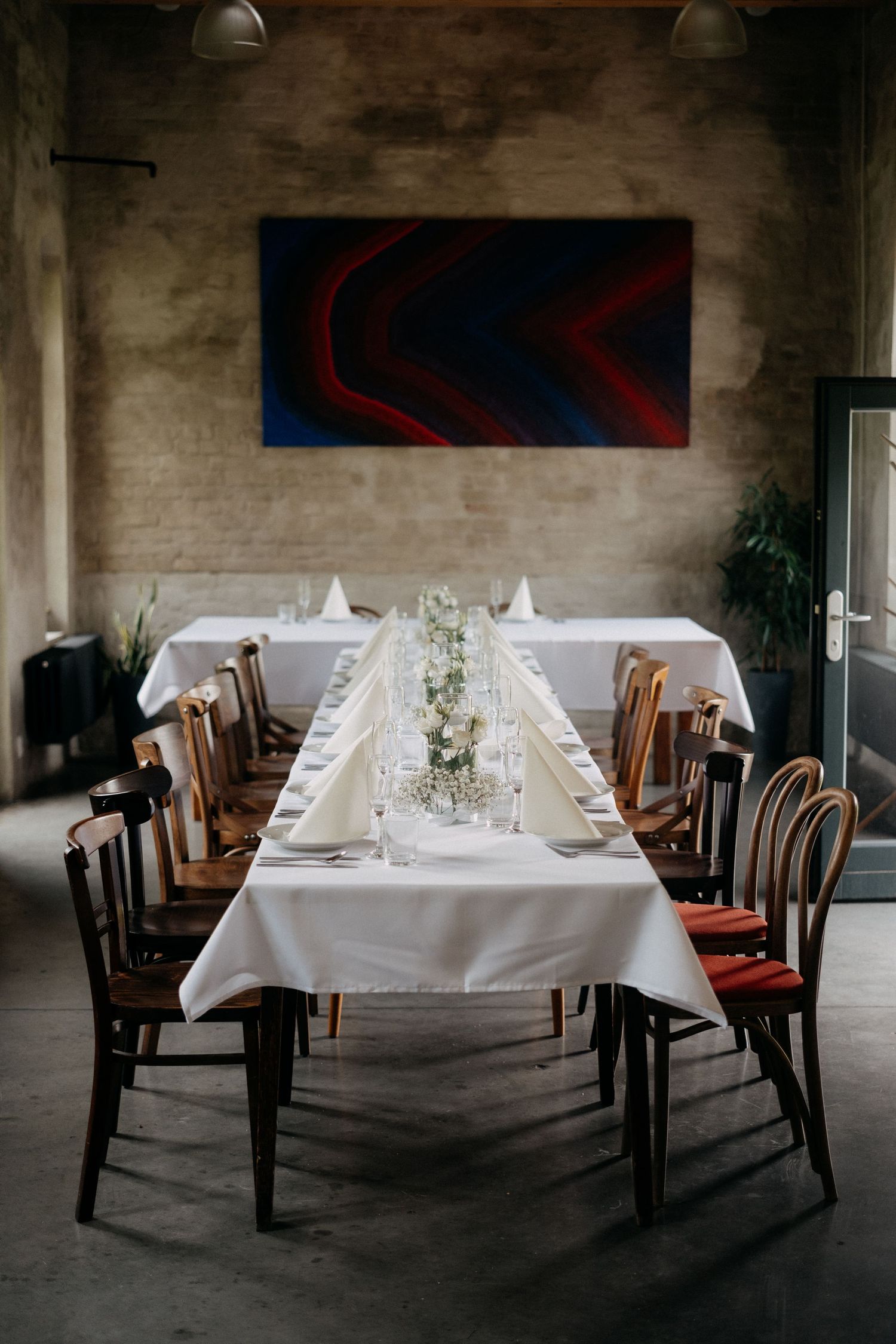 Elegant dining table setup with white linens and wooden chairs beneath modern red and blue wall art.