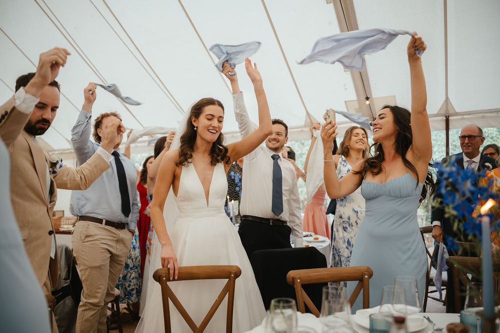 Wedding guests throw paper airplanes at a joyful celebration inside a white tent reception venue.