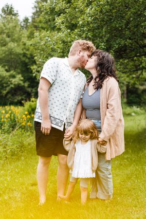 A family shares tender moments together in a sunny meadow with wildflowers and green trees in the background.