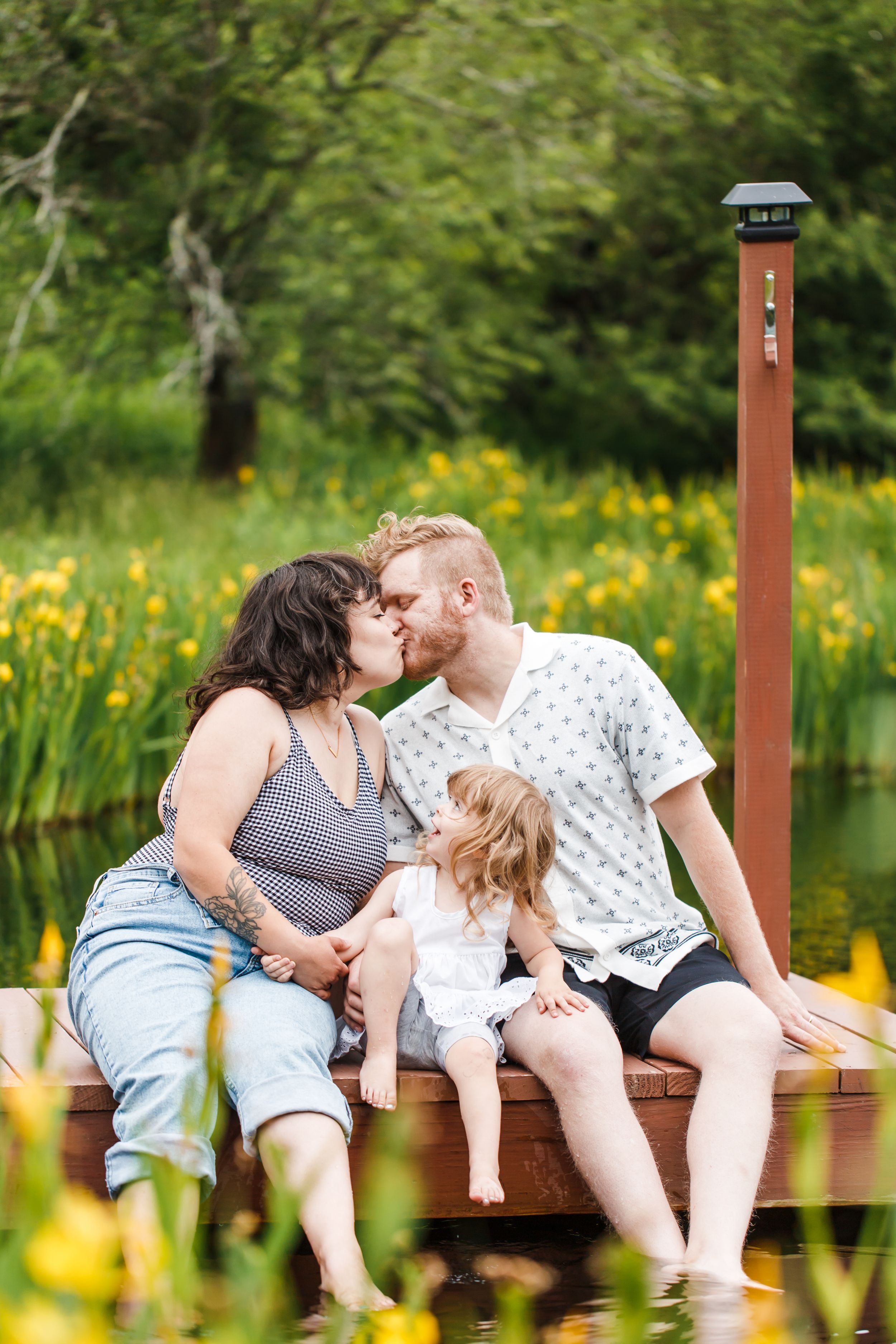 A family relaxes together on a wooden dock surrounded by yellow wildflowers and a peaceful pond.