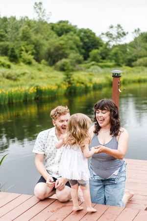 A joyful family moment captured on a wooden dock by a pond surrounded by lush greenery and yellow wildflowers.