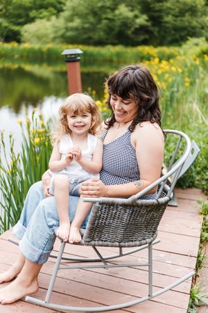 A peaceful lakeside scene with people relaxing in a wicker rocking chair on a wooden dock surrounded by yellow flowers.
