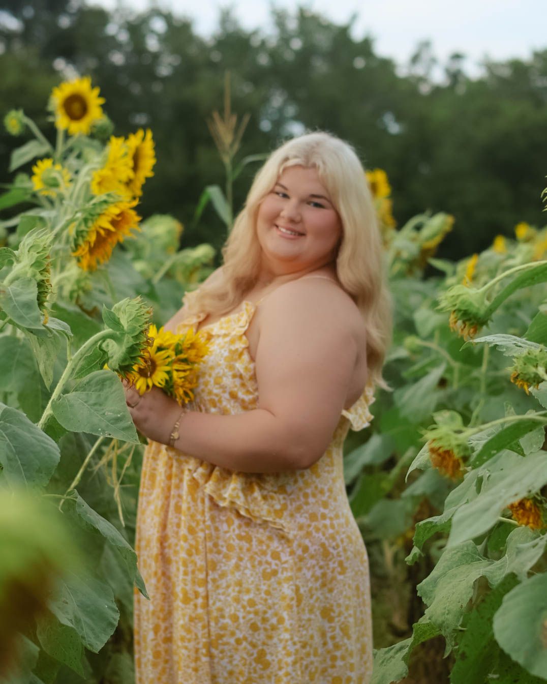 A person in a yellow floral dress poses cheerfully among tall sunflowers.