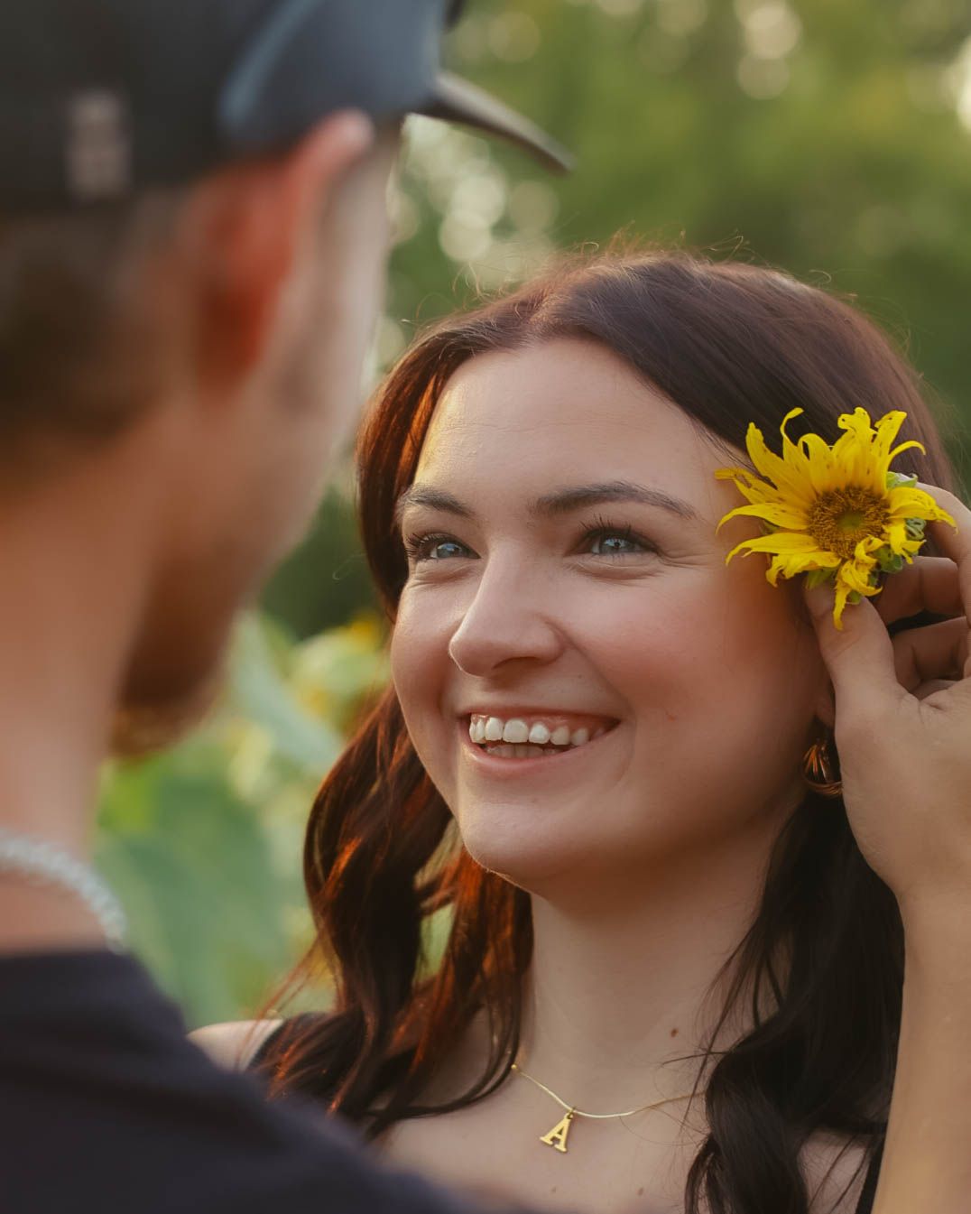 A person smiling while someone places a sunflower in their hair during a photoshoot.