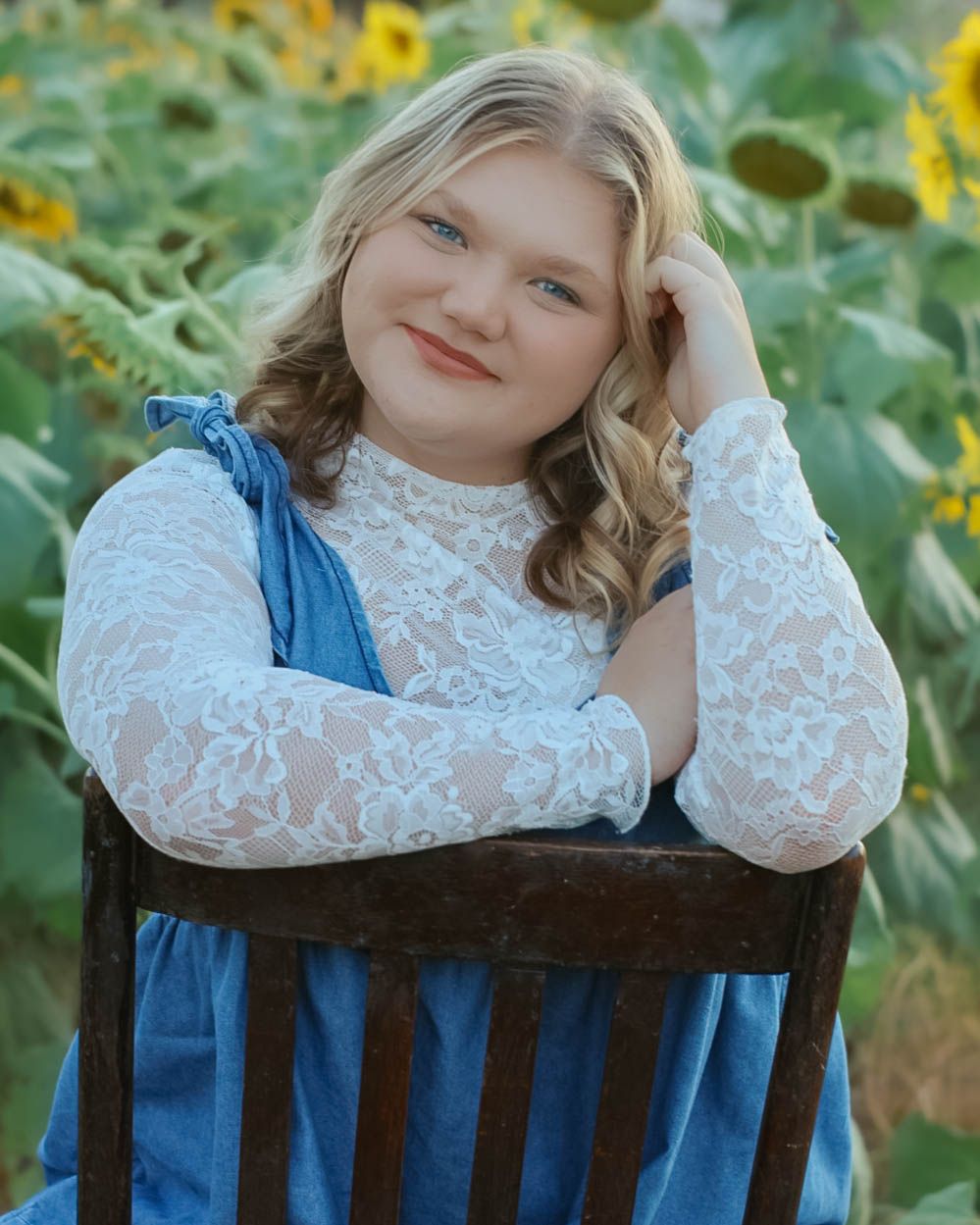 A person in a blue dress and white lace top poses on a wooden chair in a sunflower field.