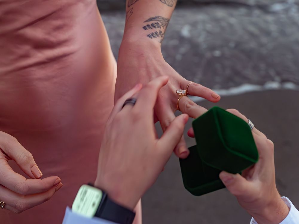 Someone in pink holds a green velvet ring box during a beach proposal.