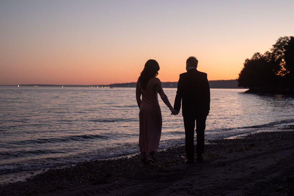 Silhouetted couple holding hands on beach at sunset with orange sky reflecting on calm water.