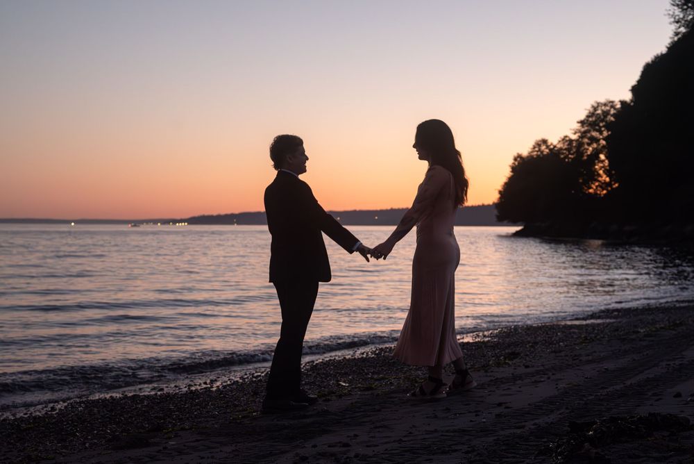 Two silhouettes hold hands on a beach at sunset with water and trees in the background.
