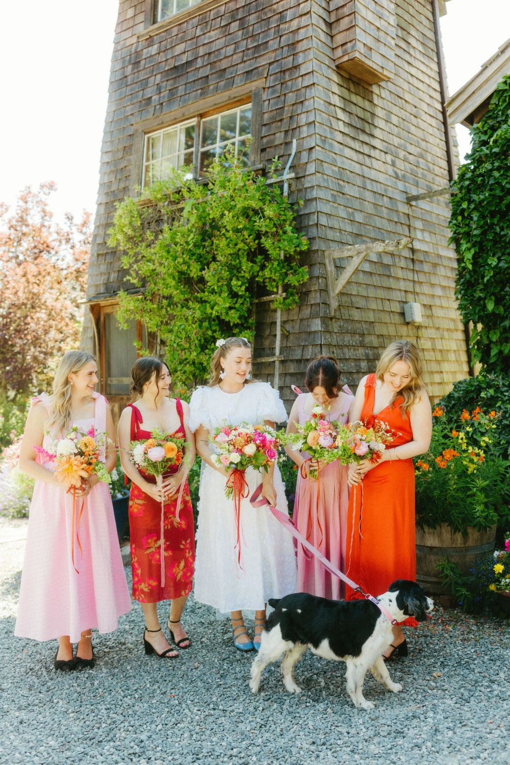 Wedding party in pink and coral dresses gathered outside stone building with garden backdrop.